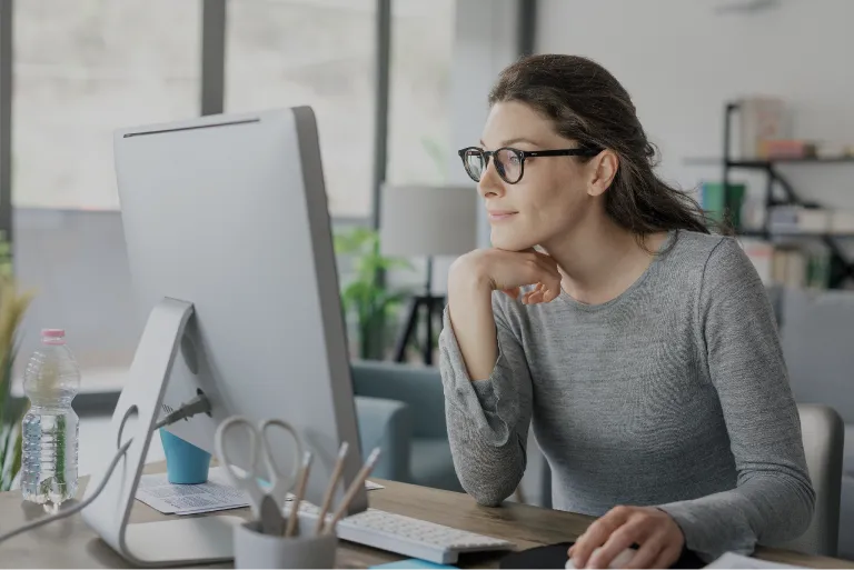 A woman wearing glasses looks at her computer screen while sitting at a desk