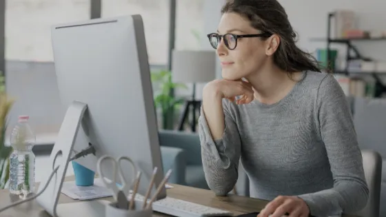 A woman wearing glasses looks at her computer screen while sitting at a desk 